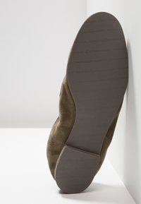 Semelle d'une chaussure en daim olive à enfiler reposant contre un coin blanc, embossée du logo « Kennel & Schmenger » sur la semelle en caoutchouc.