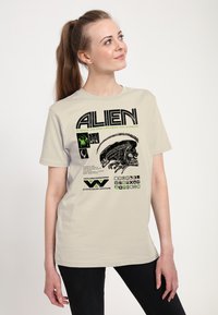 Beżowy t-shirt z czarno-zielonym nadrukiem graficznym zawierającym słowo "ALIEN", ilustrację stworzenia oraz różne symbole i tekst.