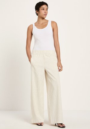 Woman standing with hands in pockets wearing a white sleeveless top and cream wide-leg pants with thin black stripes.