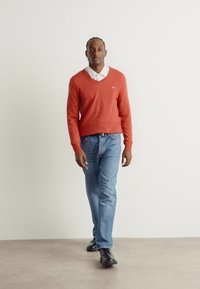 Un pull rouge à col en V porté sur une chemise blanche à col, associé à un jean bleu clair et des chaussures noires. Fond uni.