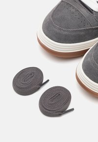 Embouts de baskets en daim gris avec semelles intermédiaires blanches et semelles extérieures marron à côté de deux lacets gris enroulés sur fond blanc.
