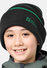 Glunderend kind met een zwarte gebreide muts met groene strepen en logo, en een grijze zip-up jack tegen een plain achtergrond.