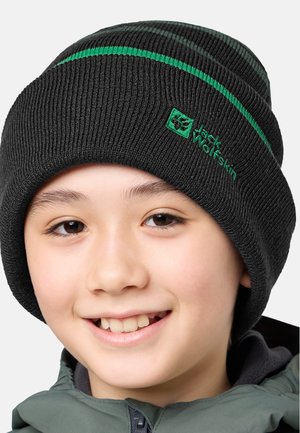 Glunderend kind met een zwarte gebreide muts met groene strepen en logo, en een grijze zip-up jack tegen een plain achtergrond.