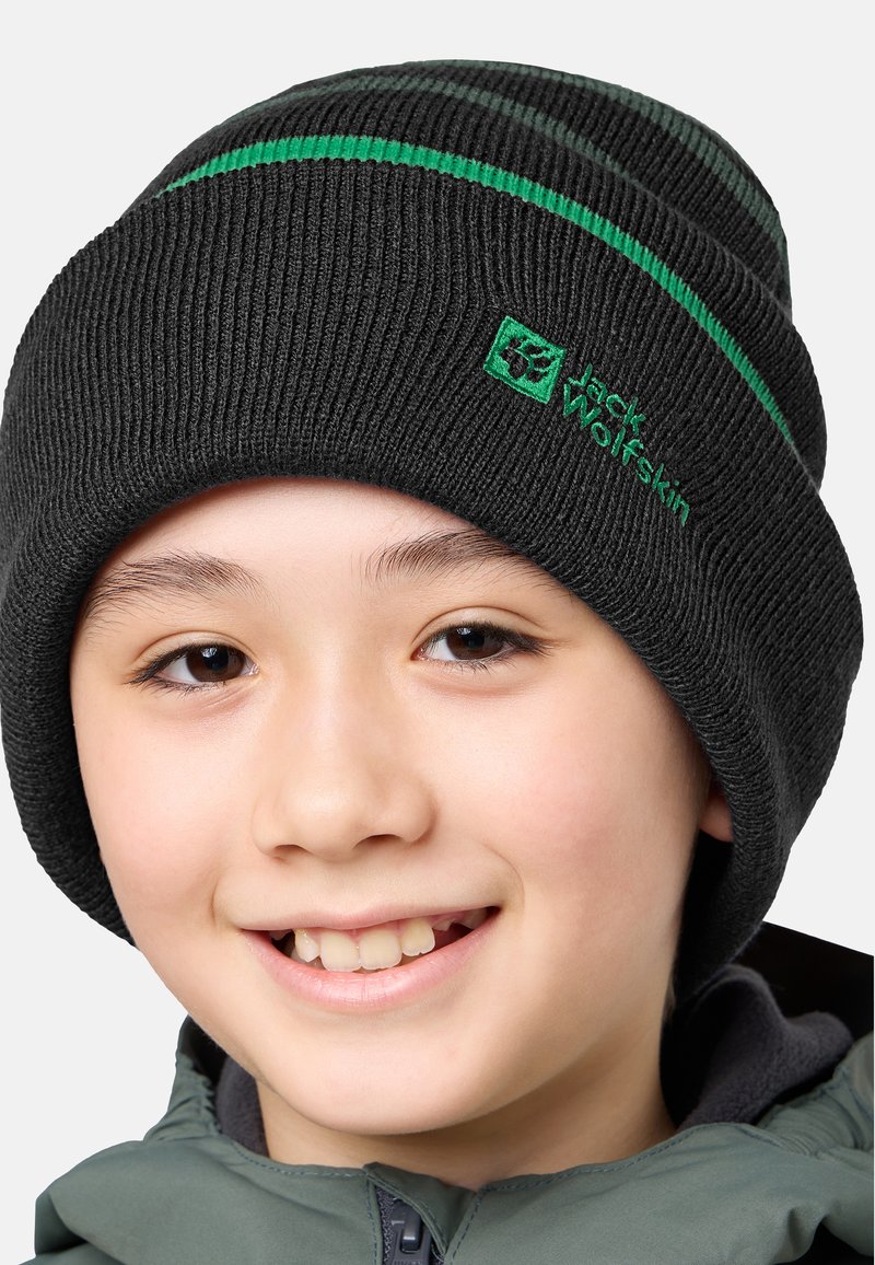 Glunderend kind met een zwarte gebreide muts met groene strepen en logo, en een grijze zip-up jack tegen een plain achtergrond.