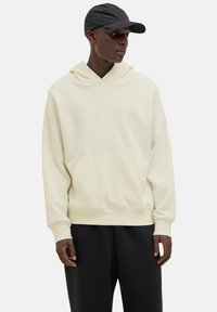 Cremefarvet hoodie med kengurulomme, ribbede manchetter og en afslappet pasform, parret med sorte sweatpants og en sort cap.