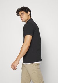 Juodas polo marškinėliai iš medvilnės, trumpomis rankovėmis ir audiniu apdirbtu apykakle, derinami su šviesiai smėlinėmis kelnėmis. Paprastas dizainas, be matomų raštų.