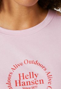 Camiseta de algodón rosa claro con texto circular en rojo. El texto incluye "Helly Hansen" y "Desde 1877" con elementos decorativos. Textura suave.