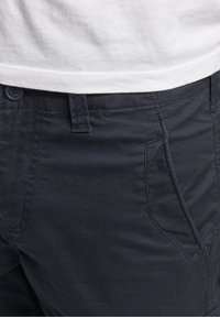 Gros plan sur une personne portant un pantalon bleu marine foncé avec une poche avant et une chemise blanche rentrée.