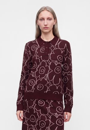 Vrouw met lang blond haar die een bordeauxkleurige top met lange mouwen en een rok draagt met grote lichtroze bloemenpatronen.