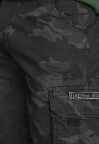 Pantaloni cargo in motivo camouflage nero, realizzati in cotone, con una tasca laterale e una patch con la scritta "ALPHA INO." Tessuto testurizzato.