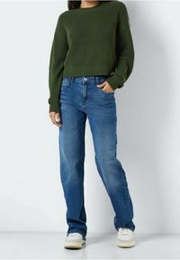 Pull vert en tricot à manches longues, associé à un jean bleu à jambes droites et des baskets blanches. La tenue présente une coupe décontractée et un matériau texturé.
