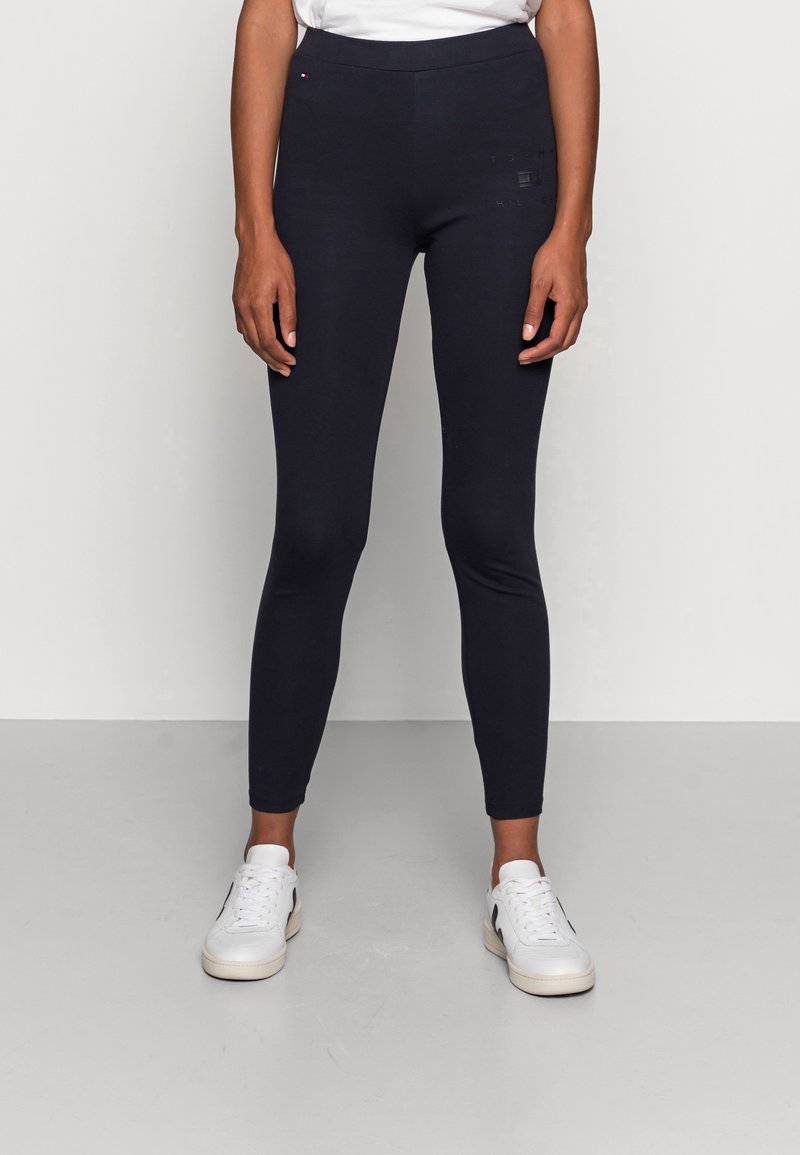 Personne portant un legging noir ajusté et des baskets blanches, se tenant contre un fond gris clair uni.
