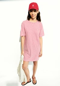 Rood-wit gestreepte korte mouwen jurk, casual ontwerp, gecombineerd met een rode pet en zwarte slippers, met een lichtgrijze jas in de hand.