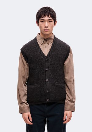 Un homme porte un gilet en maille sombre boutonné sur une chemise beige à rayures et un pantalon sombre, devant un fond clair et uni.