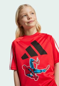Crvena sportska majica s velikim crnim Adidas logom i šarenom Spider-Man grafikom. Kratki rukavi s bijelim detaljima sa strane.