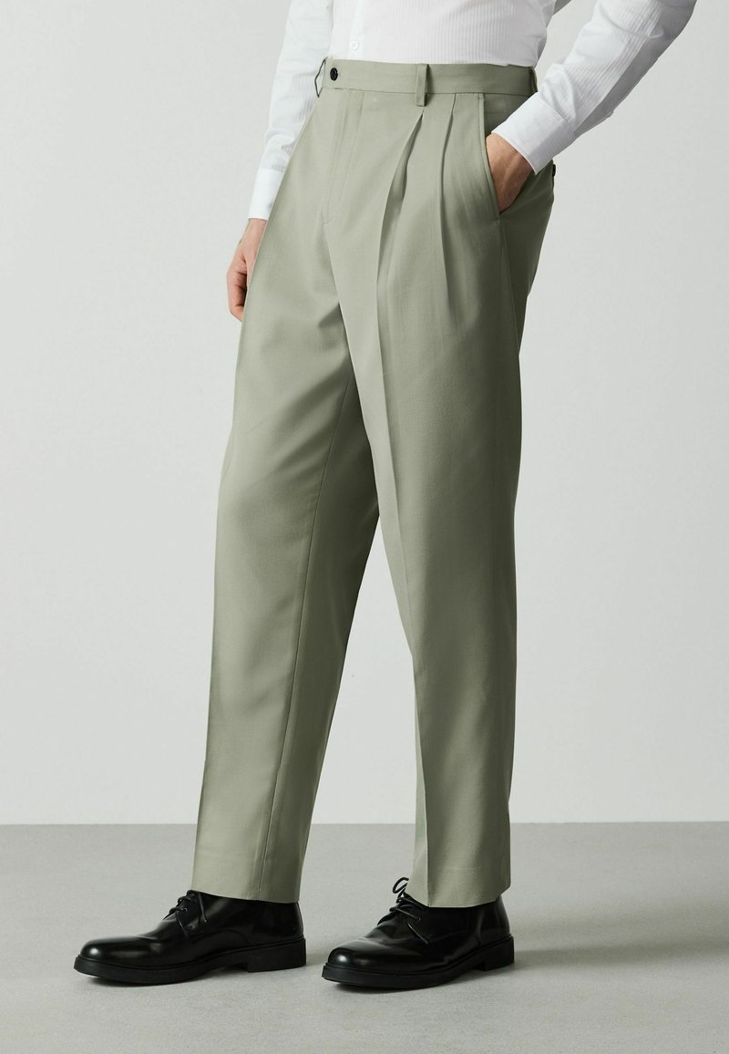 Homme portant un pantalon plissé vert olive clair, une chemise blanche et des chaussures habillées en cuir noir, debout sur un sol gris.