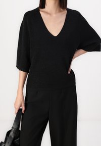 Femme portant un haut noir à col en V et manches courtes ainsi qu'un pantalon noir, tenant un sac à main noir avec une main sur la hanche.