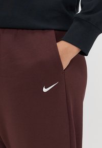 Bordové tepláky so bočnou vreckom, s bielym logom Nike Swoosh na stehne a mäkkou textúrou látky.