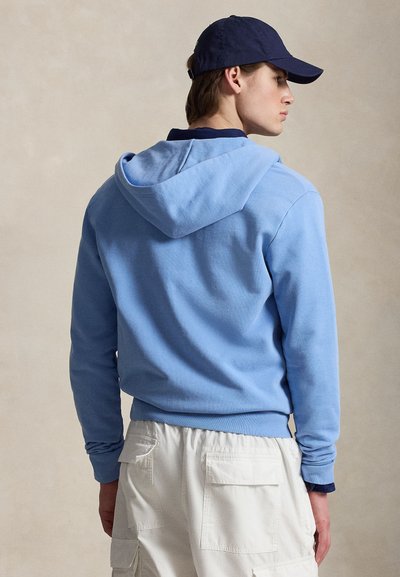 Jauns vīrietis, ģērbies gaiši zilā kapučjakā, baltās cargo biksēs un tumši zilā beisbola cepurē, skatoties no sāniem pret vienkāršu fonu.