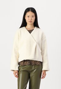 Kremfarget fleecejakke med omslagsdesign og vide ermer, lagdelt over en topp med leopardmønster, kombinert med olivengrønne bukser.