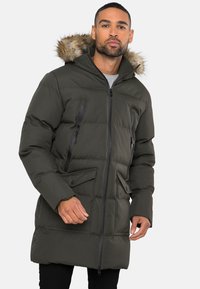 Gewatteerde parka jas in donkergroen, met een met bont gevoerde capuchon, ritszakken op de borst en knoopzakken aan de voorkant. Zachte, gewatteerde textuur.