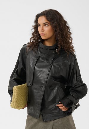 Frau mit lockigem Haar, die eine schwarze Lederjacke trägt und eine große geflochtene beige Clutch hält, schaut zur Seite.