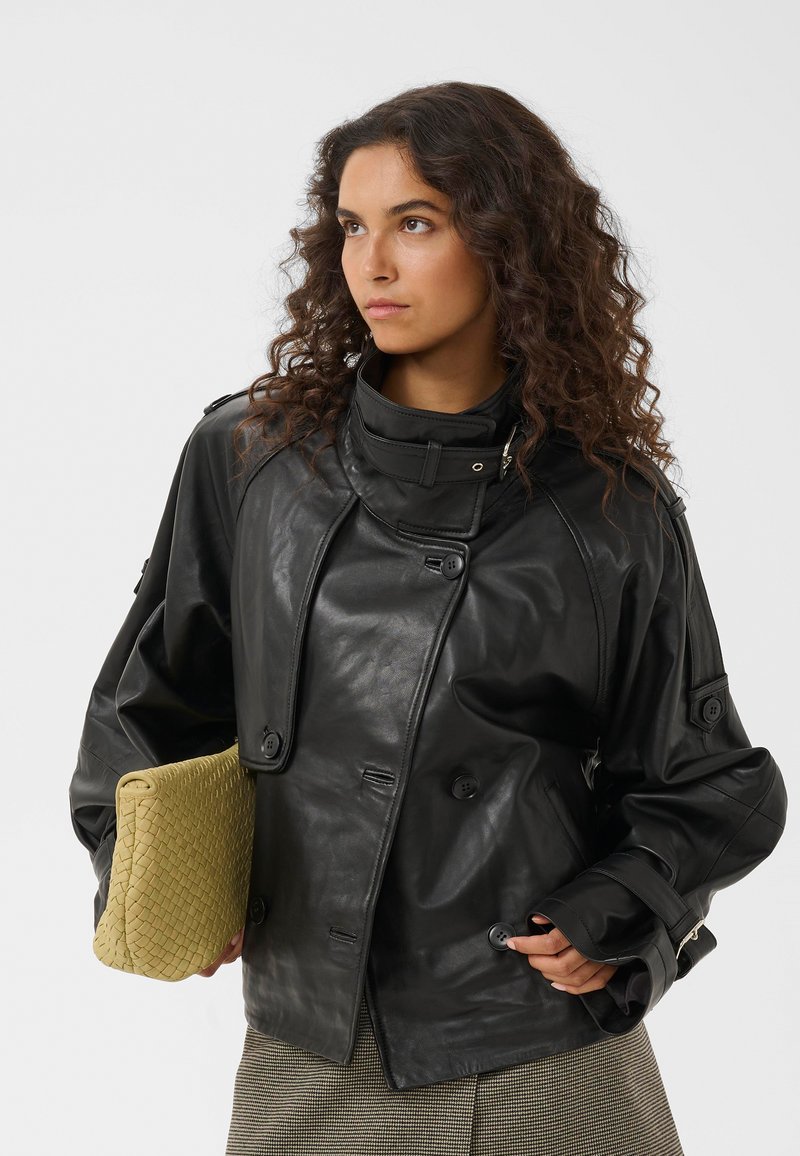 Femme aux cheveux bouclés portant une veste en cuir noire et tenant une grande pochette beige tissée, regardant sur le côté.