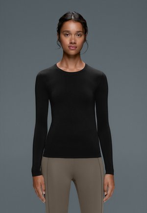 Femme aux cheveux foncés tirés en arrière, vêtue d'un haut noir ajusté à manches longues et d'un pantalon marron clair sur un fond gris uni.