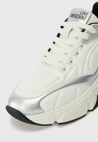 Sapatilha branca com detalhes prateados brilhantes, texturizados na parte superior, atacadores planos e uma sola de borracha preta grossa. Apresenta a marca "LOVE MOSCHINO".