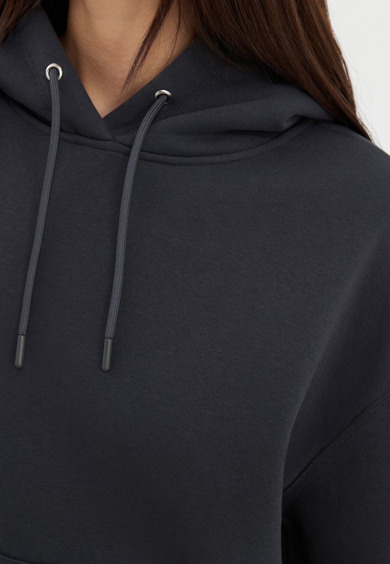 Ciemnoszara bluza z kapturem o fakturowanej tkaninie, z kapturem ze sznurkiem, srebrnymi oczkami i detalami w szyciu. Zbliżenie na dekolt.