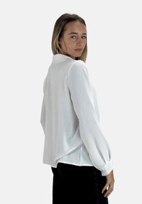 Witte blouse met lange mouwen en een subtiele textuurafwerking, voorzien van een kraag en luchtige mouwen, weergegeven tegen een effen achtergrond.
