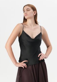 Model poseert zelfverzekerd, gekleed in een zwart satijnen top met dunne bandjes en een vloeiende bordeauxrode rok, met een effen witte achtergrond.
