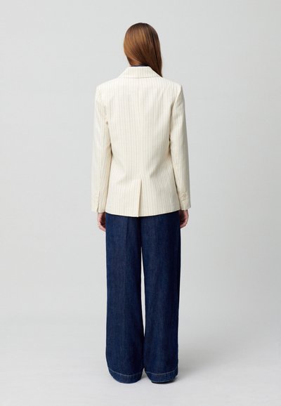 Blazer beige a righe con maniche lunghe, spacco singolo sul retro, abbinato a pantaloni in denim scuri a gamba larga, dettaglio orlo grezzo.