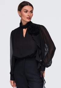 Femme portant un chemisier noir à col montant avec des manches bouffantes transparentes et une grande fleur en tissu sur une épaule, associé à un pantalon noir.