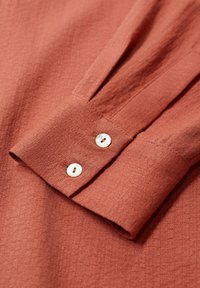 Manche de chemise couleur rouille avec deux boutons blancs sur le poignet, montrant un tissu texturé et des plis cousus près du poignet.