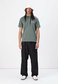 Green cotton t-shirt with a graphic design on the chest, paired with loose-fitting black pants and white sneakers. White bucket hat worn.