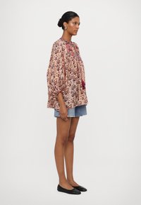 Bluse mit floralem Muster und Raffung, heller rosa Hintergrund und roten Akzenten, kombiniert mit Jeansshorts. Schwarze Ballerinas.