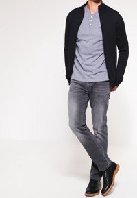 Svart zip-up cardigan över en grå henley-tshirt, parat med grå jeans och svarta läderstövlar med tan-färgade sulor. Avslappnad outfit.