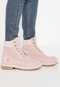 Personne portant des bottines à lacets roses clairs avec des cols rembourrés et des semelles beiges, associées à un jean en denim bleu.