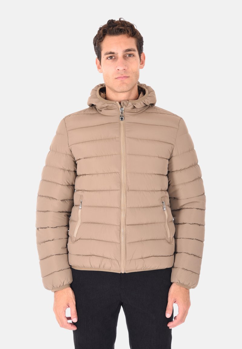 Uomo con capelli corti e ricci, che indossa un giubbotto imbottito beige con cappuccio e cerniera frontale, in piedi davanti a uno sfondo chiaro e semplice.