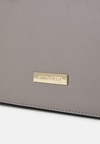 Sac en synthétique gris avec une surface texturée, une plaque logo dorée et des coutures le long des bords.