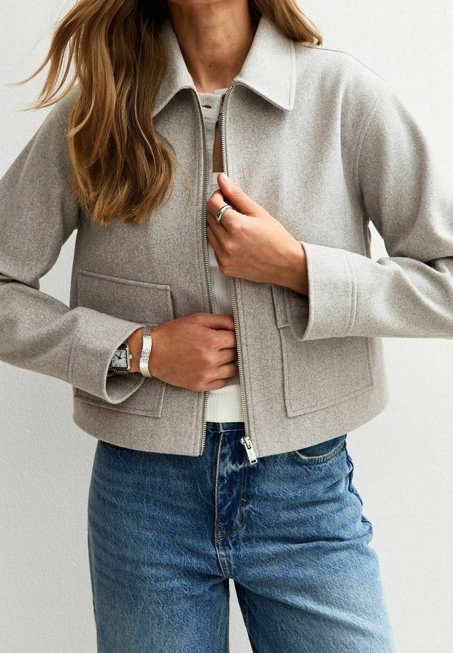 Veste grise claire cropped avec col, poches avant et fermeture éclair ; associée à un jean en denim bleu. Texture lisse, design minimaliste.