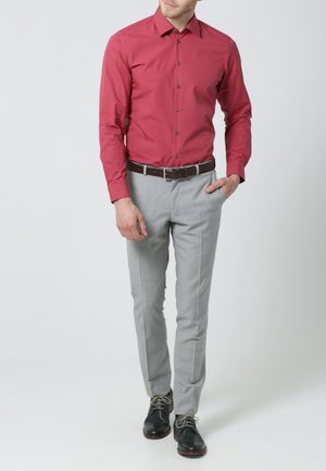 Rød tætsiddende skjorte med lange ærmer, skjortekrave med knapper, kombineret med lysegrå skræddersyede bukser og sorte lædersko.