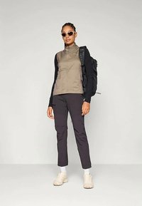 Μπεζ γιλέκο με φερμουάρ πάνω από μαύρο πουκάμισο με μακρύ μανίκι, συνδυασμένο με σκούρο παντελόνι και λευκά αθλητικά παπούτσια. Τα αξεσουάρ περιλαμβάνουν γυαλιά ηλίου και μαύρο σακίδιο.