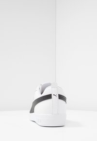Weißer Sneaker mit einem strukturierten schwarzen Streifen, glattem Lederfinish, flacher weißer Sohle und einem Logo auf der Rückseite. Minimalistisches Design.
