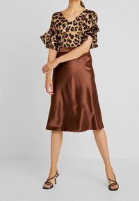 Blouse à imprimé animal avec des manches volantées, associée à une jupe midi en satin marron. Le modèle porte des talons aiguilles noirs à lanières.