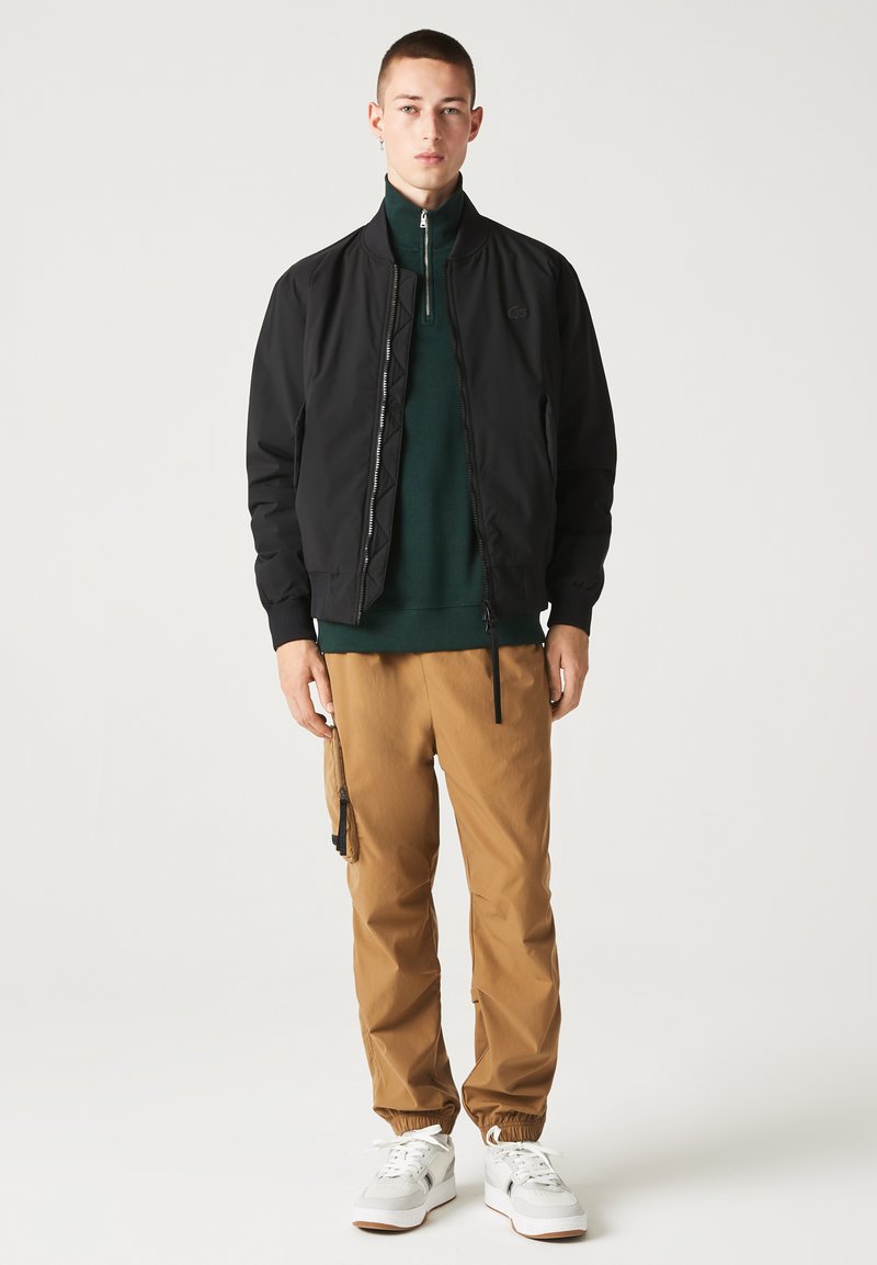 Veste bomber noire avec fermeture éclair, associée à un pull à col zippé vert et à un pantalon de jogging beige avec une taille ajustable et des poches.