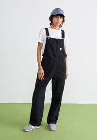 Schwarze Denim-Overalls mit verstellbaren Trägern, frontaler Tasche und Logo-Patch, kombiniert mit einem weißen T-Shirt und grauen Schuhen auf grünem Boden.