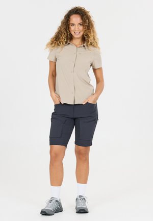 Femme souriante aux cheveux bouclés, les mains dans les poches, portant une chemise beige à manches courtes, un short cargo foncé, des chaussettes blanches et des baskets grises.