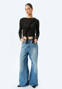 Schwarzes Langarmoberteil mit Rüschendetails, kombiniert mit hellblauen weit geschnittenen Jeans und schwarzen spitzen Schuhen. Glatte Stoffbeschaffenheit.